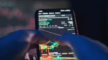 Karanlık bir odadaki akıllı telefon ekranında şifreli para çizelgelerini analiz eden ellerin yakın çekimi. Finans ve teknoloji projeleri için ideal.