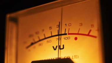 Işıldayan analog VU metre yakın plan ve iğnesi yüksek. Ilık retro ışıklandırma, ses mühendisliği, müzik stüdyoları veya analog teknoloji temaları için ideal olan klasik cazibesini ön plana çıkarıyor.