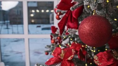 Karla kaplı bir Noel ağacında parlak kırmızı bir takı asılı duruyor. Yumuşak arka plandaki pencereden görülebilen karlı bir kış manzarası var. Tatil temaları için ideal.