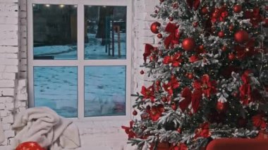 Geniş bir pencereden karla kaplı bir açık hava manzarası ve kırmızı süslemeler, noktalar ve sıcak ışıklarla süslenmiş bir Noel ağacı içeren neşeli bir tatil sahnesi. Dekorasyon tatil havasını çağrıştırıyor.