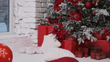 Süslü bir Noel ağacı, kırmızı süslemeler ve fiyonklarla, paketlenmiş hediyelerin arasına yerleşmiş, sıcak, şenlikli bir atmosfer yaratıyor. Tatil projeleri ve kutlamalar için mükemmel..