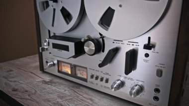 Play levyesini retro-to-reel teyp kayıt cihazında çalıştıran bir elin yakın çekimi. Dönen manyetik makaralar ve klasik analog kontroller zamansız ses ekipmanlarının cazibesini vurgular.