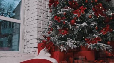 Çok güzel dekore edilmiş kırmızı süslemeler, noktalar ve parıldayan ışıklarla süslenmiş bir Noel ağacı. Sahnede kırmızı hediye kutuları, beyaz tuğlalı duvarlar ve karlı bir ortam var.