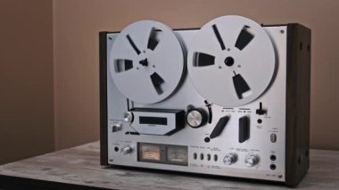 Yakın çekim retro bobin ses kaydedicisi, dönen bant makaraları, detaylı düğmeler ve VU sayaçları. Ahşap bir masaya yerleştirilmiş, yüzyılın ortalarından kalma bir cazibe yayıyor, müzik severler ve klasik koleksiyoncular için ideal.
