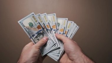 Düz kahverengi bir arka plan karşısında 100 dolarlık banknotların hayranı ellerini sayıyor. Konsept: finans, gelir, ödemeler ve kişisel refah yönetimi.