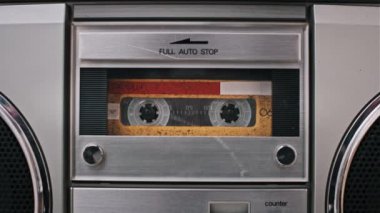 Bir kaset açık bir retro stereo desteye yerleştirilir, ardından kompartıman kapatılır ve çalma başlatılır. Turuncu etiketli banta ve güverte mekaniğine odaklan..