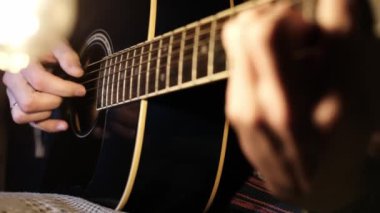 Bir müzisyenin elleri, parlayan bir ampulün yumuşak ışığı altında telleri tıngırdatırken, siyah bir gitara odaklanmış bir gitar üzerinde ayrıntılı bir eylem sergiliyor..