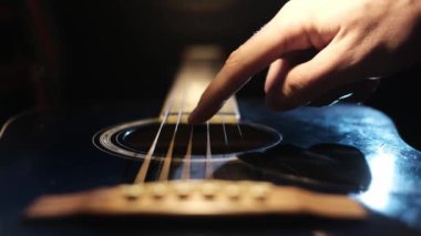 Gitar tellerine hafifçe basan bir parmak yakın plan, loş, odaklanmış stüdyo ışıkları altında karanlık bir gitar gövdesi üzerinde bir eylem gerçekleştiriyor ön planda köprü ile.