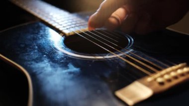Bir el, gitar telleri üzerinden ritmik hareketi yakalayan, dokulu vücudu ve dairesel açılışı vurgulayan siyah bir akustik gitar çalar..