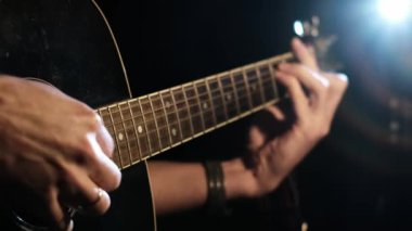 Koyu renkli bir akustik gitar çalan biri. Odaklanılan parmaklar fretboard 'da ve eller tıngırdıyor, hepsi siyah arka plan ve parlak ışık merceğine karşı..