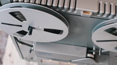 Eski bir makaradan makaraya kasetçaların yakın çekim görüntüsü ve iki makaranın çalma sırasında dönmesi. Gümüş bant makaraları ve gri oyuncu genel bir perspektiften görüntülenir, dönüşe odaklanırlar.