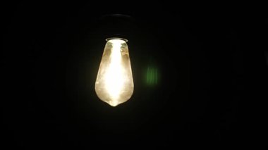 Karanlık bir odada asılı parlayan bir Edison ampulüne kısaca dokunan bir elin yakın çekimi karmaşık iplik ve sıcak ışık detaylarını gözler önüne seriyor. Ampul çok ince titreşiyor..