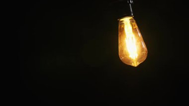 Eski bir Edison ampulü koyu bir arka planda parlıyor ve asılıyor. Detaylı iplik ve sıcak, ışıl ışıl ışığa odaklanarak karamsar ve atmosferik bir manzara yaratılıyor..