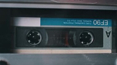 Eski bir çalgıcının içinde dönen siyah bir ses kasetinin yakın görüntüsü. Mavi etiket ve temiz bant penceresi, geçmişe dönük ses teknolojisini gösterir.