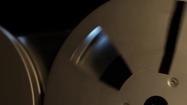 Yakın çekim, odak görünümü, her iki makaranın da ses kaydı sırasında ışık ve gölgelerin etkileşimini vurgulayan düşük ışıklandırma altında, eski bir makaradan makaraya teyp eşliğinde dönüşünü gösterir