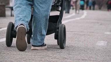 Kot pantolon ve spor ayakkabı giymiş bir kadının bacaklarının yakın çekimi, şehir ortamında bir bebek arabasının asfalt bir yolda ilerlemesi..