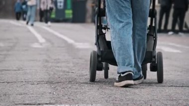 Bir annenin parkta belirlenmiş bir patikadan bebek arabasını iterken çekilmiş düşük açılı görüntüsü. Arka planda başka insanlar da görülüyor. Odak noktası anne bacakları ve bebek arabasının hareketi..