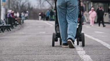 Genç bir anne, diğer park ziyaretçilerinin arasında sakin bir yürüyüşün tadını çıkararak, bir parktaki asfalt bir yolda bebek arabasını itiyor. Düşük kamera açısı bebek arabalarının hareketine ve anne ayaklarına odaklanıyor.