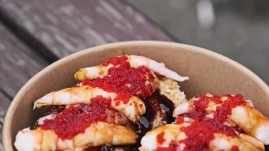 Parlak kırmızı tobiko balığı yumurtası ve ahşap bir yüzeyde duran tuzlu sosla kaplı karides suşisi dolu paket servis kasesinin detaylı görüntüsü. Japon deniz ürünleri lezzeti.