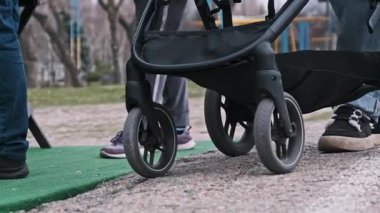 Siyah bebek arabalarının yakın plan çekimleri asfaltlı bir yüzeye itiliyor, arka planda insanların bacakları ve ayakkabıları görünüyor..