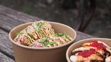 İçinde suşi burgerleri olan iki paket servis kasesinin yakın plan detayları. Çeşitli malzemelere odaklanın: kırmızı tobiko ve soslu karides ve ahşap bir yüzey üzerinde sunulan yeşil garnitürlü ton balığı..