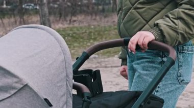 Bir anne modern bir bebek arabasının deri sapını nazikçe tutar, park ortamında hareketsiz durur, muhtemelen sakinleştirir ya da bebeğiyle ilgilenir..