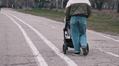 Bir kadın, parktaki asfalt yolda bebek arabasıyla yürüyor. Kamera bacaklarına ve bebek arabasının tekerleklerine odaklanıyor. Arka planda diğer park ziyaretçileri ve ağaçlar görülüyor