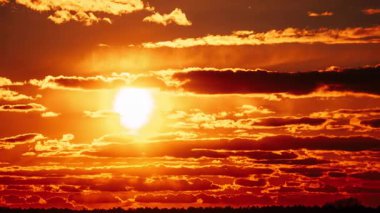 Ateşli turuncu bir gün batımının zamanı. Güneş dramatik, karanlık, katmanlı bulutların arasından, siluetli bir ormanın üzerinde parlıyor..