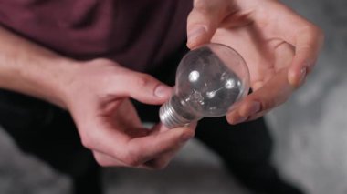 Erkek ellerinin tozlu ve geleneksel bir akkor ampulü tutarken ve çevirirken yukarıdan aşağıya yakın görüntüsü, filamanı inceler ve gri bir arkaplana karşı tabanı vidalayarak.