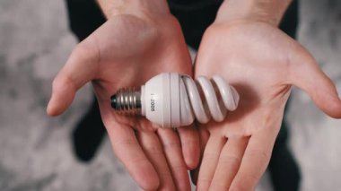 Erkek ellerinin nazikçe tuttuğu ve kameraya doğru enerji tasarrufu sağlayan spiral bir CFL ampulü sunduğu yakın çekim. Hafifçe yakınlaştırıyorum. Enerji korunumu ve modern ışıklandırmaya odaklanın.