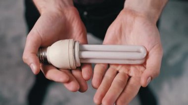 Bir adam elindeki beyaz floresan lambayı yakından inceleyerek cam tüplerin ve tabanının durumunu belirlemek için çeviriyor. Yapısal detaylara ve enerji tasarrufu tasarımına odaklan.