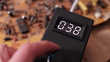 Kontrol düğmesini çeviren bir elin yakın görüntüsü dijital ekrandaki sayıları artırıyor. Bir atölyede elektronik test ekipmanındaki rakamlarla temsil edilen elektrik voltaj değişimini gösterir.