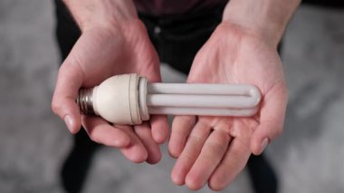 Işıklandırma cihazını gösteren bulanık arkaplana karşı CFL 'in enerji tasarruflu florasan ampulünü dikkatle tutan bir kişinin yukarıdan aşağı görüntüsü.