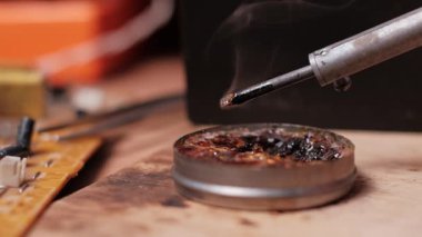 Ağır çekimde, kızgın bir lehim ucunun reçine akışının üzerinde uçarken sigara içerken görüntüsü. Elektronik tamir ekipmanları ve devre kartı ahşap bir çalışma tezgahında görülebilir.