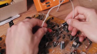Tozlu bir elektronik devre kartındaki arızaları teşhis etmek için kırmızı ve siyah çoklu metre sondalar kullanan ellerin yakın çekimi. Ahşap tezgahın arka planında turuncu bir çokölçer görünür..