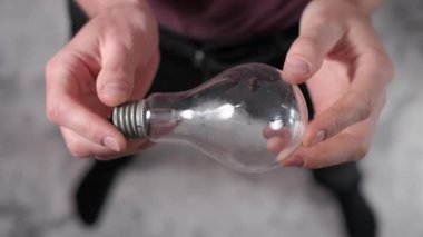 Tepedeki perspektif, erkek elleri dikkatlice yakalıyor. Geleneksel parlak ampulü döndürerek cam yapısını, filamanını ve metal vida tabanını gösteriyor. Teknik inceleme odağı.