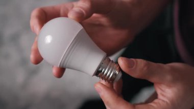 Beyaz enerji tasarruflu bir LED ampulü tutan ve döndüren erkeklerin yakın çekim görüntüleri. Buzlu kubbeye odaklan ve temelleri vidala, modern aydınlatma teknolojisini ve sürdürülebilir enerji kullanımını betimle.