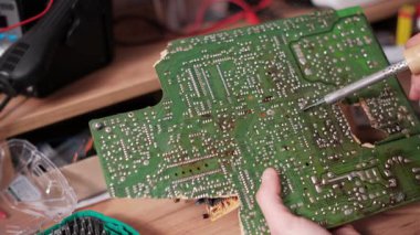 Hasarlı yeşil devre kartına lehimleme bileşenlerini titizlikle kapatırken dağınık bir elektronik tamir atölyesi ortamında kızgın ütü kullanarak hassas çalışma odaklı....