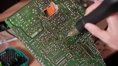 Bir elektronik tamir işlemi sırasında, yeşil baskılı devre kartının ters tarafındaki lehimleme noktalarına sıcak hava uygulayan bir teknisyenin eli yakın çekim görüntüsü.