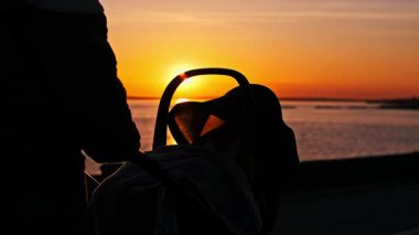 Gün batımında deniz kenarında yürüyen genç bir annenin silueti bebek arabasını itiyor. Sıcak altın ışık suya yansıyarak, bir aile yürüyüşü için huzurlu bir akşam atmosferi yaratır..