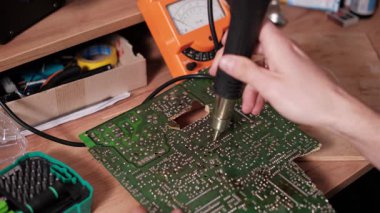 Yakın çekim elektronik teknisyeni, yeşil baskılı PCB devre kartının arkasındaki lehimleri ısıtmak ve sökmek için sıcak hava istasyonu aracı kullanıyor.