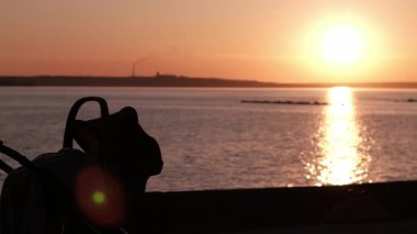 Canlı bir günbatımında bebek arabasıyla sahil yolunda yürüyen ve iten bir annenin yan görünüş silueti. Çocuğuyla akşam gezintisi yaparken güneş suya yansıyor..