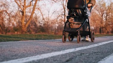 Düşük açılı izleme çekimi siyah bebek arabasının tekerleklerini yuvarlamaya ve anne ayaklarının asfalt yolda yürümesine odaklanır. Ebeveyn altın saat batımında bebek arabasını kenara itiyor.