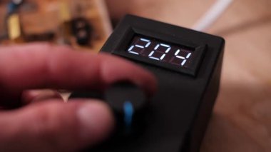 Elektronik kontrol ünitesi veya güç kaynağının dijital ekranında hızla değişen, muhtemelen voltaj veya sıcaklığı ayarlamak için döndürme düğmesini çeviren bir elin yakın çekim görüntüsü.