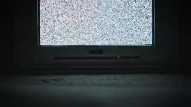 Klasik CRT televizyonun düşük açılı görüntüsü titreyen statik gürültü, ekranda beyaz kar deseni. Karanlık odada ekranın altında kırmızı ışık parlıyor. Sinyal yok, analog parazit konsepti.