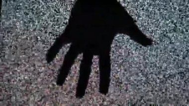 Yakından çekilen görüntü, çerçevenin tepesinden yavaşça beliren siluetli bir eli yakalar, parmaklarını uzatır ve titreşen siyah beyaz TV parazitinin arka planına doğru hareket eder..