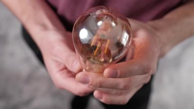 Yüksek açılı yakın plan, kehribar renkli yuvarlak bir cam eldiven tutan erkek ellerini gösteriyor. Edison tarzı LED filaman ampulü, eski dünya estetiğini ve enerji tasarruflu filamanların yapısını vurguluyor..