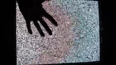Koyu renk siluet eli, renkli parazit gösteren eski bir televizyonun ekranına uzanıp nazikçe dokunuyor. El, kumlu ve canlı yüzey boyunca yavaşça hareket eder.