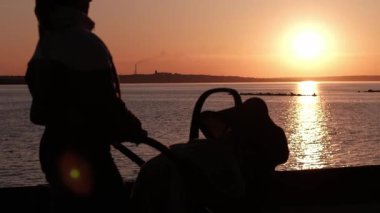 Bir annenin bebek arabasıyla rıhtımda durup gün batımını izlediği siluet. Parlak güneş sakin suyu yansıtıyor, uzak kıyı manzaralı huzurlu bir akşam manzarası yaratıyor..