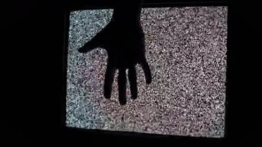 Siluetli el yavaşça iniyor ve eski bir CRT televizyonunun titreşen statik ekranına dokunuyor. Karanlık oda ortamında analog beyaz gürültü karışıklığı gösteren ürkütücü bir korku konsepti.